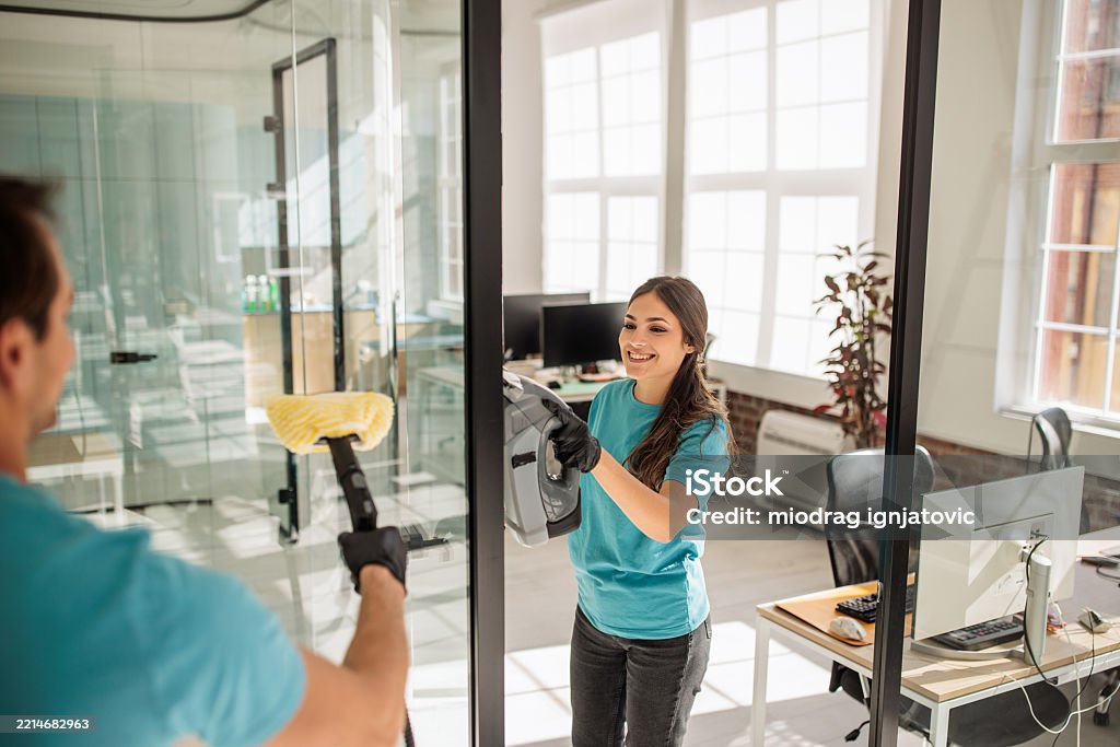 Office cleaners in blue uniforms and gloves clean glass walls in a bright, modern workspace with desks and computers—showcasing a spotless, professional business environment.