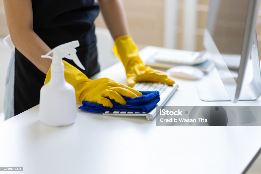 Office cleaner disinfecting computer keyboard with microfiber cloth and eco-friendly spray. Commercial cleaning service for workplace hygiene and sanitized office surfaces.